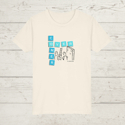 Beige t-shirt with a graphic design 'Overcomer, 1 John 5:4' on a wooden surface with cartoon and scrabble tiles