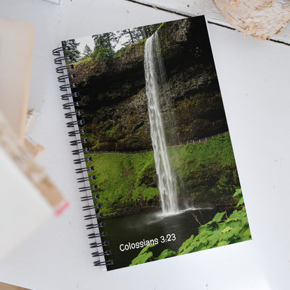 Notebook with a waterfall design and 'Colossians 3:23' text on a white surface.