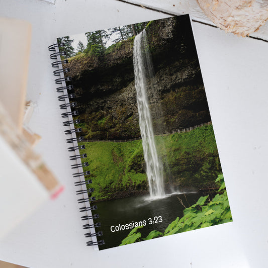 Notebook with a waterfall design and 'Colossians 3:23' text on a white surface.