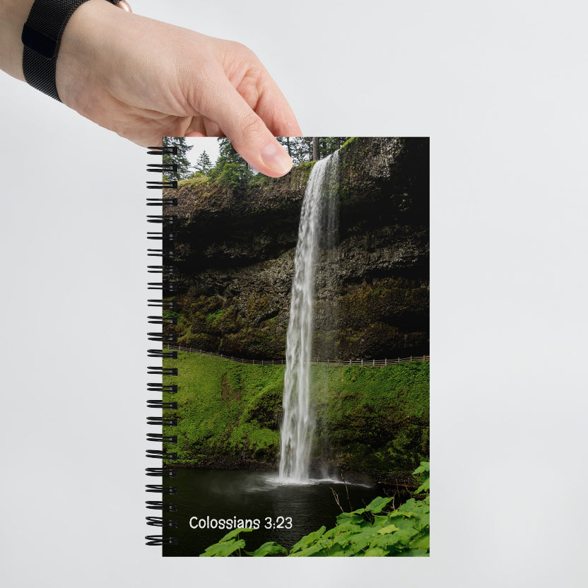 Notebook with a waterfall design and 'Colossians 3:23' text, held by a hand.