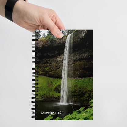 Notebook with a waterfall design and 'Colossians 3:23' text, held by a hand.