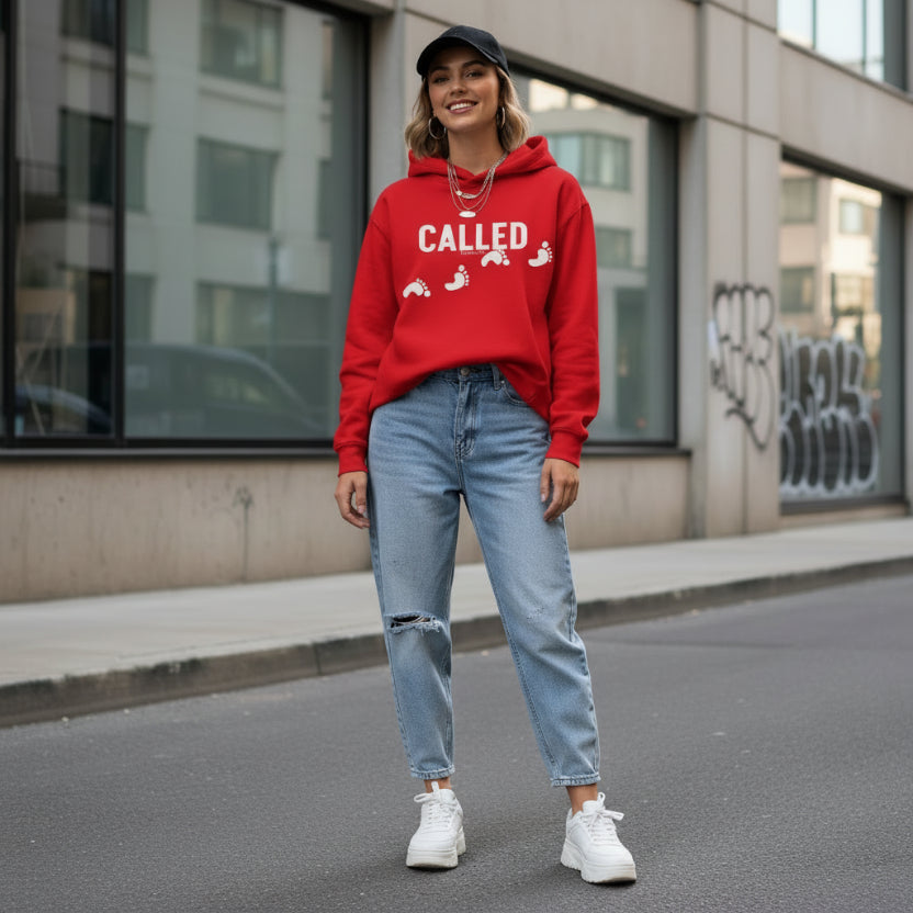 Woman wearing a red 'Called' hoodie with text, blue jeans, and white sneakers on a city street.