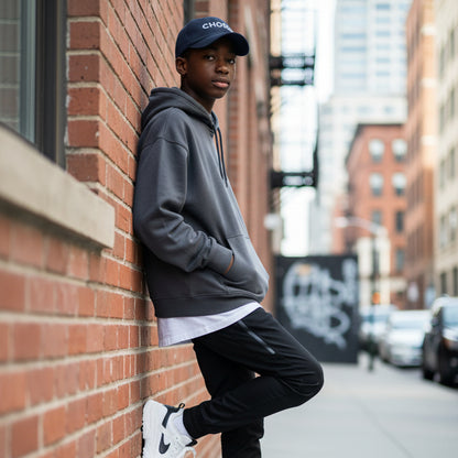 Black cap with 'Chosen' embroidered, teen leaning against building in the city