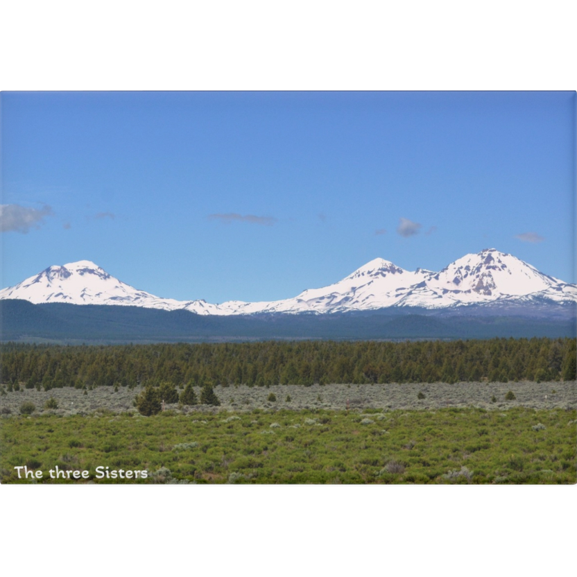 Three snow-capped mountains with a clear blue sky and green landscape in the foreground.