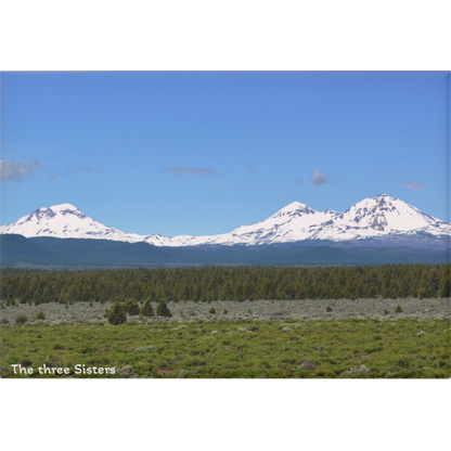 Three snow-capped mountains with a clear blue sky and green landscape in the foreground.