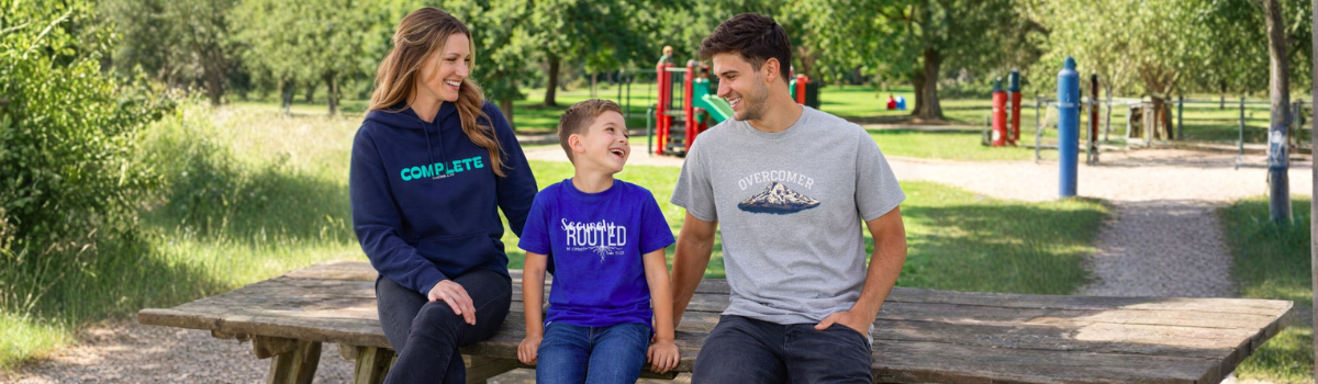 Family of three sitting on a picnic bench at a park portraying original brand design apparel