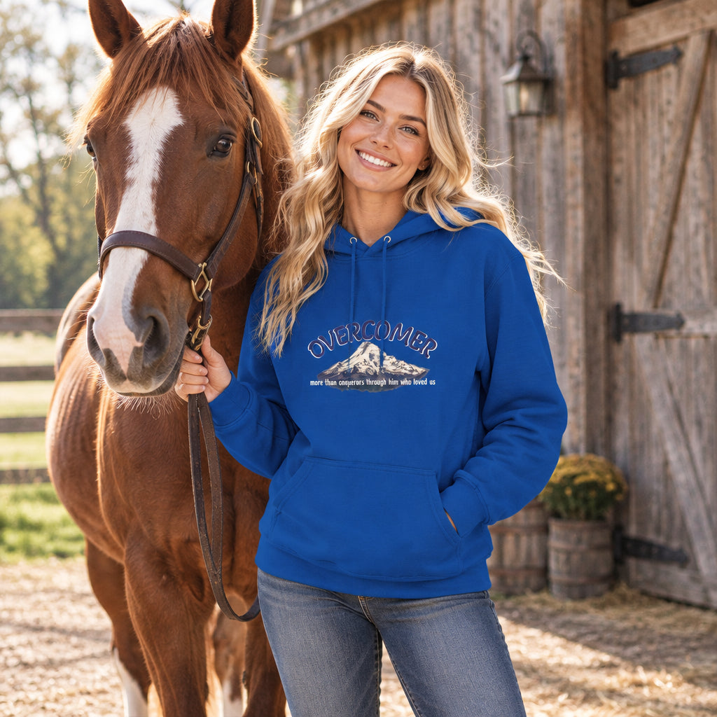 A woman stands outdoors beside a brown horse, wearing a bright blue hoodie with a mountain graphic and the word 'OVERCOMER' on the front. The background shows a rustic wooden barn.