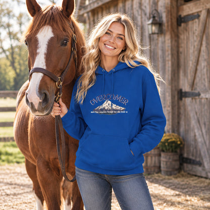 A woman stands outdoors beside a brown horse, wearing a bright blue hoodie with a mountain graphic and the word 'OVERCOMER' on the front. The background shows a rustic wooden barn.