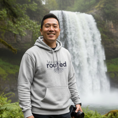 Gray hoodie with text 'Securely Rooted' near waterfall holding a camera