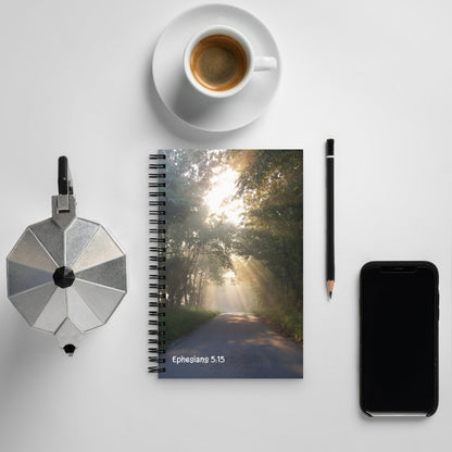 Notebook with a scenic cover, coffee cup, pen, phone, and metallic object on a light surface.