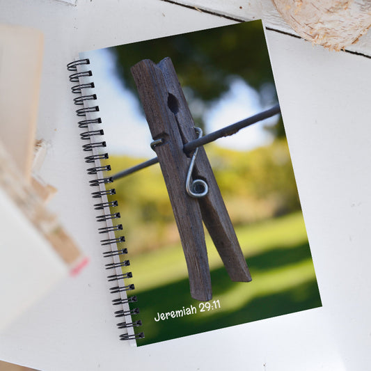 Notebook with a clothespin on a string design and 'Jeremiah 29:11' text, placed on a wooden surface.