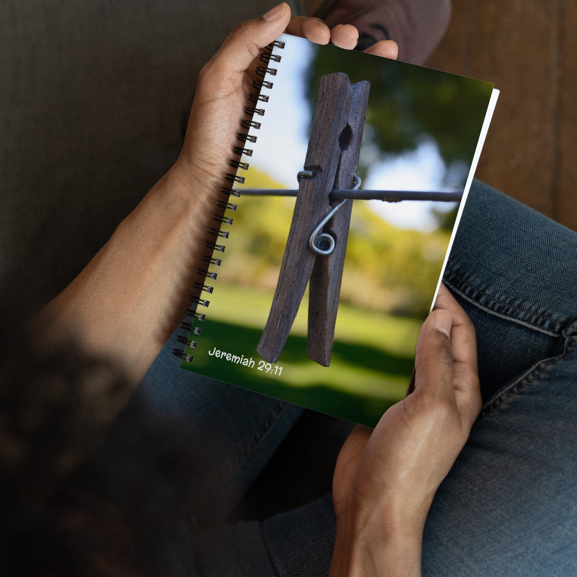 Person holding a notebook with a wooden clothespin and 'Jeremiah 29:11' text on a blurred natural background